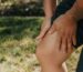 A close-up image of a person holding their knee, indicating pain or injury, with greenery in the background.
